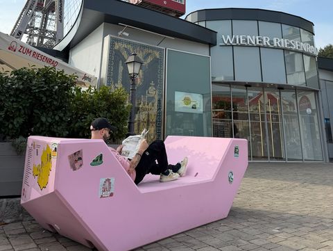 Eine Person liest auf einem rosa Enzi vor dem Wiener Riesenrad im Wiener Prater eine Zeitung.