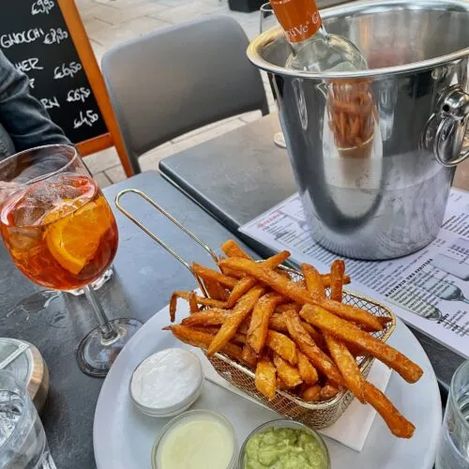 On a table is a plate of sweet potato fries and three different sauces in small bowls. Next to it is an Aperol Spritz with ice cubes and a glass of water.