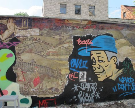 Mural showing a historical landscape and a large cartoon figure wearing a blue cap with graffiti text in the foreground.