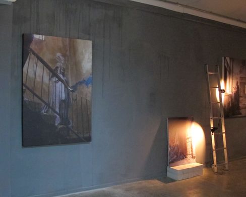 Dimly lit room with three pictures hanging on the wall and a ladder with a lamp casting light on one picture.
