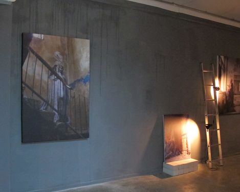Dimly lit room with three pictures hanging on the wall and a ladder with a lamp casting light on one picture.