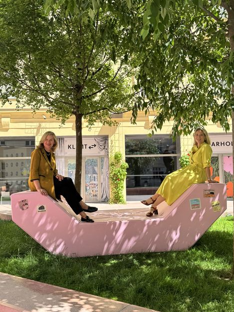 Two women sitting on a pink Enzi seating sculpture in the green courtyard of MuseumsQuartier Wien in front of exhibition posters.