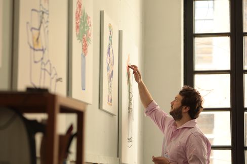 Man in pink shirt looking at and touching a drawing on a wall with several framed pictures next to a window.