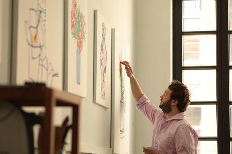 Man in pink shirt looking at and touching a drawing on a wall with several framed pictures next to a window.