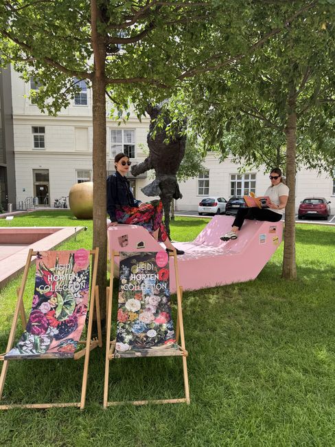 Zwei Frauen sitzen auf einem rosa Enzi-Sitzmöbel im MuseumsQuartier Wien unter schattigen Bäumen. Im Vordergrund stehen Liegestühle der Heidi Horten Collection, im Hintergrund eine Skulptur und die begrünte Parkanlage.