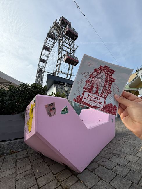 Ein Wiener-Riesenrad-Sticker wird vor dem Riesenrad und einem rosa Enzi in die Kamera gehalten.