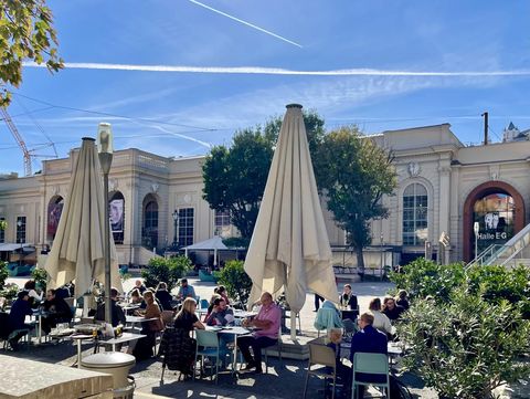 Mehrere Menschen sitzen an Tischen unter großen Sonnenschirmen auf einer Terrasse vor einem historischen Gebäude.