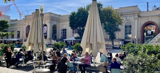 Several people are sitting at tables under large parasols on a terrace in front of a historic building.