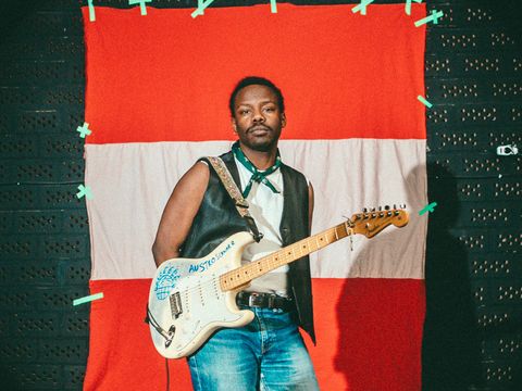 Mann mit schwarzer Lederweste, weißem Hemd und Jeans steht vor rot-weiß-rotem Hintergrund und hält eine E-Gitarre mit Aufschrift.