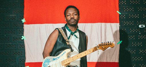 Mann mit schwarzer Lederweste, weißem Hemd und Jeans steht vor rot-weiß-rotem Hintergrund und hält eine E-Gitarre mit Aufschrift.