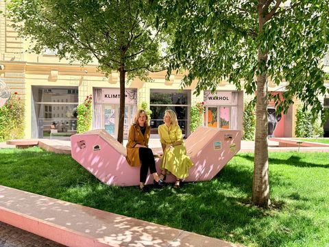 Two women sitting on a pink Enzi seating sculpture in the green courtyard of MuseumsQuartier Wien in front of exhibition posters.