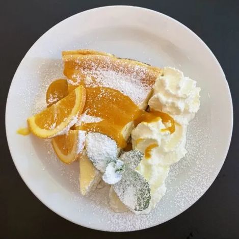 Fresh cake on a white plate garnished with whipped cream, orange cream, powdered sugar, and orange slices.