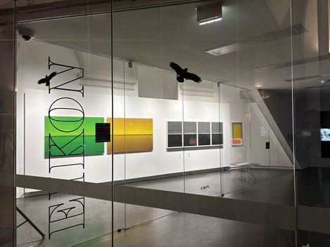 Blick durch eine Glaswand mit der Aufschrift "EIKON" auf eine Galerie mit mehreren rechteckigen, farbigen Kunstwerken an der Wand und zwei schwarzen Vogel-Silhouetten auf der Glasfläche.