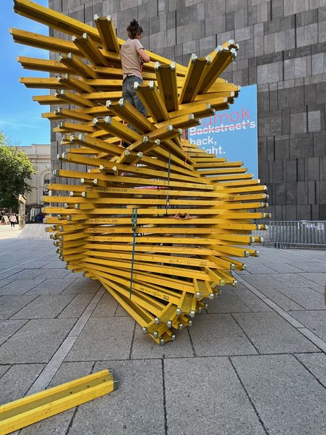 Person steht auf einer großen, spiralförmigen Skulptur aus gelben Holzlatten auf einem gepflasterten Platz vor einem Gebäude mit grauer Fassade