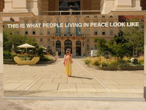 Spiegelnde Installation mit der Aufschrift 'THIS IS WHAT PEOPLE LIVING IN PEACE LOOK LIKE' vor einem historischen Gebäude, eine Person steht davor und macht ein Foto.