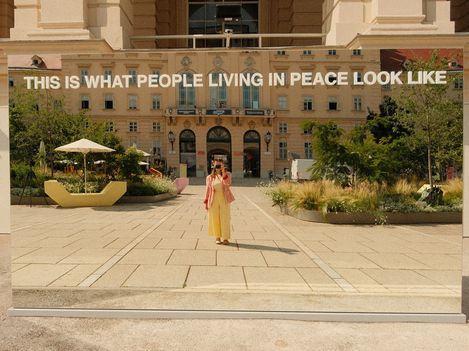 Spiegelnde Installation mit der Aufschrift 'THIS IS WHAT PEOPLE LIVING IN PEACE LOOK LIKE' vor einem historischen Gebäude, eine Person steht davor und macht ein Foto.