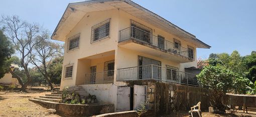 It is a bright house. In the background is a blue sky. To the left and right of the house, you can see trees and bushes. The ground in front of it is loamy.