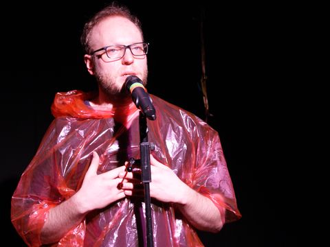 An actor is standing in front of a microphone, wearing a red, see-through rain cape.