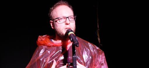 An actor is standing in front of a microphone, wearing a red, see-through rain cape.