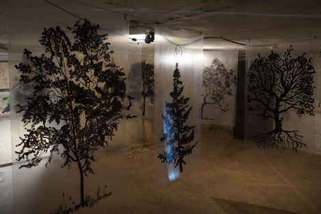 Several transparent sheets with black-and-white tree drawings hang from the ceiling in a dimly lit room.