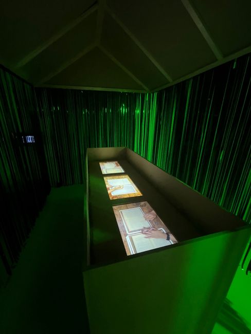 Dark room with green lighting and a long table displaying three screens projecting hands and paper.