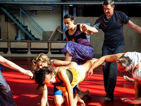 Gruppe von Menschen in einem Raum mit rotem Boden, die sich in einer choreografierten Bewegung gegenseitig stützen