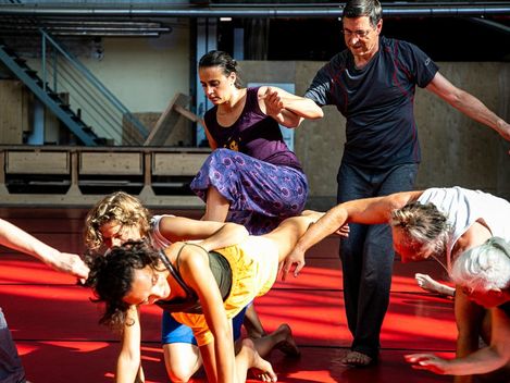 Gruppe von Menschen in einem Raum mit rotem Boden, die sich in einer choreografierten Bewegung gegenseitig stützen