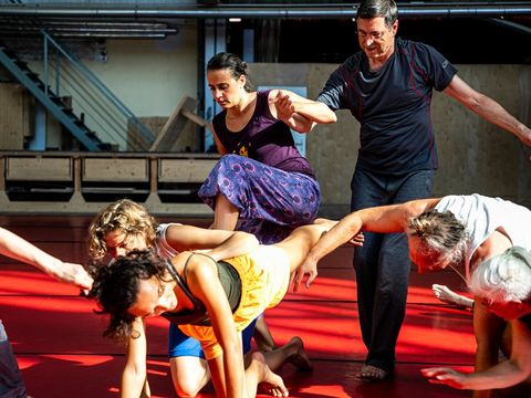 Gruppe von Menschen in einem Raum mit rotem Boden, die sich in einer choreografierten Bewegung gegenseitig stützen