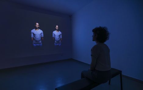 Person sitting on a bench in a dark room watching a wall projection of two identically dressed people in light shirts and dark pants.