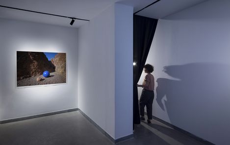 Gallery room with a photograph of a blue sphere in a rocky landscape on the wall and a person pulling aside a black curtain.