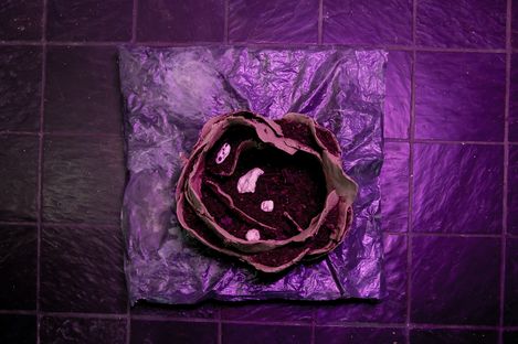 Skulptur aus geschichteten, unregelmäßig geformten Materialien auf quadratischer Unterlage vor dunklem Fliesenboden mit violettem Licht.