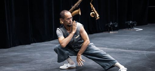 Mann in grauer Weste und Hose in tiefer Tanzpose auf Bühne mit zwei Saxophonen im Hintergrund