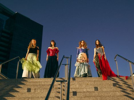 Vier Personen in fantasievollen, farbenfrohen Outfits stehen oben auf einer breiten Steintreppe. Aus der Froschperspektive aufgenommen, heben sie sich im warmen Sonnenlicht vor einem tiefblauen Himmel und einem modernen Glasgebäude ab.