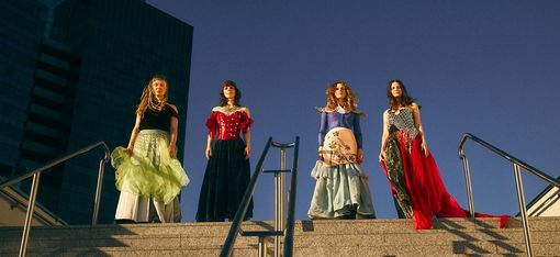 Vier Personen in fantasievollen, farbenfrohen Outfits stehen oben auf einer breiten Steintreppe. Aus der Froschperspektive aufgenommen, heben sie sich im warmen Sonnenlicht vor einem tiefblauen Himmel und einem modernen Glasgebäude ab.