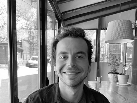 Person with dark curly hair sitting in a café with large windows and visible plant arrangement on a wooden table.