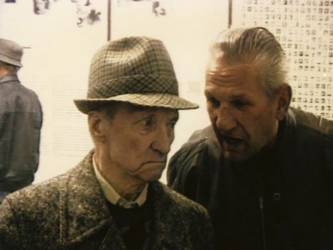 Two elderly men, one wearing a checkered hat and coat, the other with slicked-back hair, talking in front of a wall with texts and images.