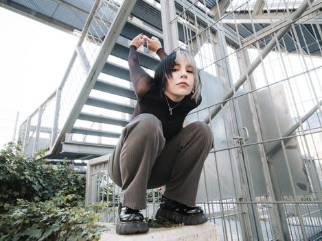 Person with black-and-white dyed hair crouches on a concrete block in front of a metal staircase holding hands behind head.