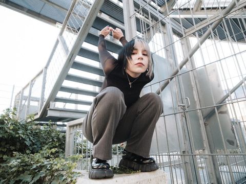 Person with black-and-white dyed hair crouches on a concrete block in front of a metal staircase holding hands behind head.