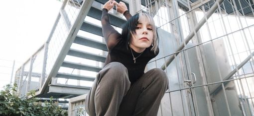 Person with black-and-white dyed hair crouches on a concrete block in front of a metal staircase holding hands behind head.
