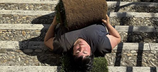 Jen Rosenblit is lying upside down on a flight of stairs, holding a piece of rolled turf in her hands