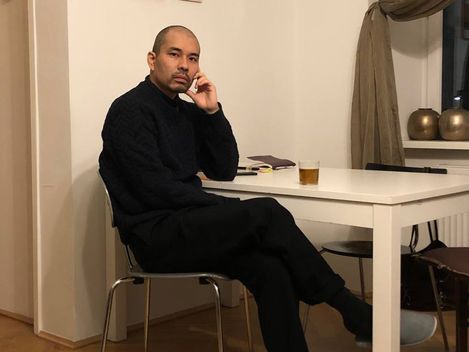 Person sitting with crossed legs at a white table in a room with parquet flooring and curtains.