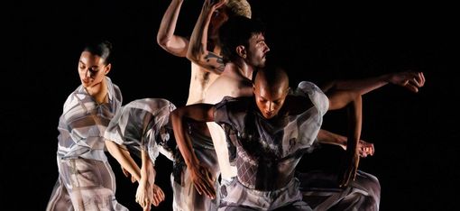 Four dancers in sheer flowing costumes move synchronously against a black background