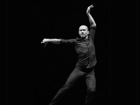 Ein Tänzer in schwarzer Kleidung führt eine ausdrucksstarke Pose mit einem Arm nach oben und dem anderen nach vorne aus vor schwarzem Hintergrund.