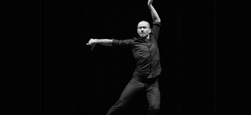 Ein Tänzer in schwarzer Kleidung führt eine ausdrucksstarke Pose mit einem Arm nach oben und dem anderen nach vorne aus vor schwarzem Hintergrund.