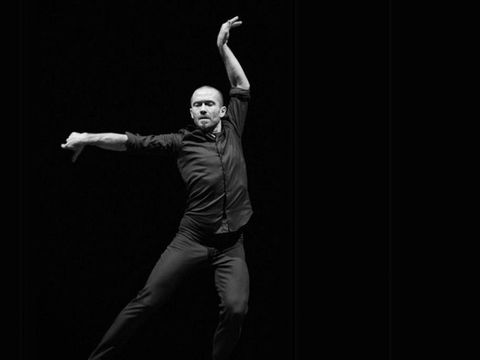 Ein Tänzer in schwarzer Kleidung führt eine ausdrucksstarke Pose mit einem Arm nach oben und dem anderen nach vorne aus vor schwarzem Hintergrund.