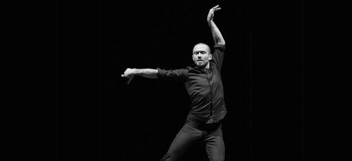 Ein Tänzer in schwarzer Kleidung führt eine ausdrucksstarke Pose mit einem Arm nach oben und dem anderen nach vorne aus vor schwarzem Hintergrund.