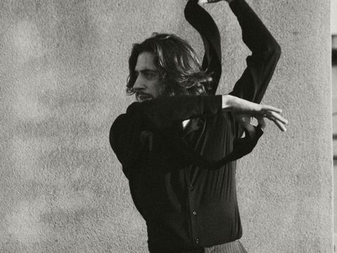 A man with shoulder-length hair wearing a dark top and striped pants dances with raised arms in front of a wall.