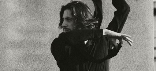 A man with shoulder-length hair wearing a dark top and striped pants dances with raised arms in front of a wall.