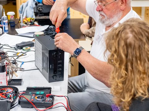 Älterer Mann mit Brille schraubt an einem schwarzen Computergehäuse an einem Tisch, während eine jüngere Person mit lockigem Haar daneben sitzt und zuschaut