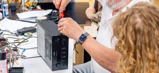 Älterer Mann mit Brille schraubt an einem schwarzen Computergehäuse an einem Tisch, während eine jüngere Person mit lockigem Haar daneben sitzt und zuschaut